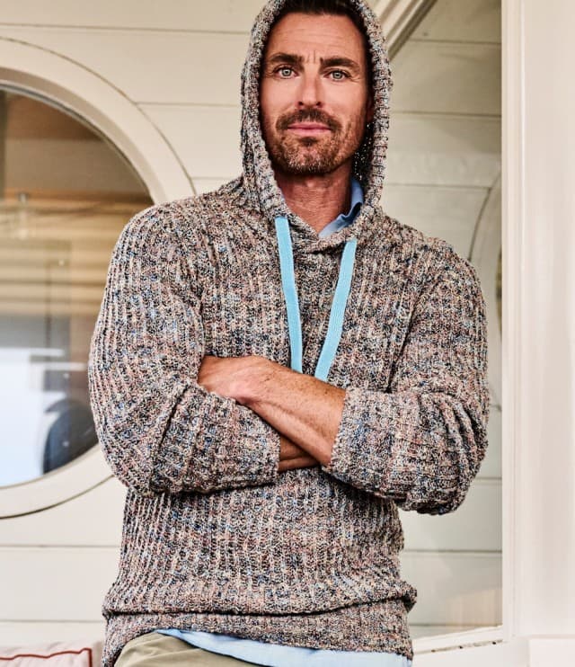 Ein Mann trägt einen grob gestrickten Strickpullover und steht mit verschränkten Armen vor einer Schiffswand.