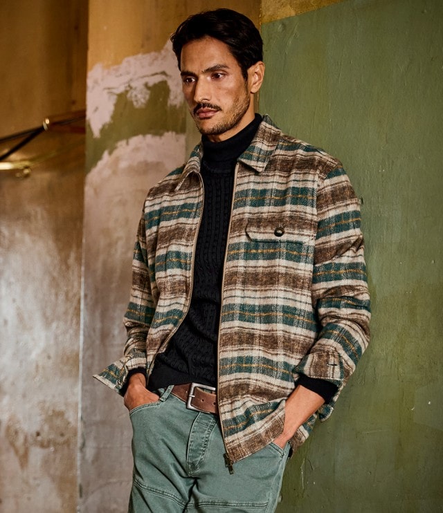 Ein Mann steht mit den Händen in den Taschen vor einer grün-braunen Wand und trägt ein kariertes Overshirt in Braun- und Grüntönen über einem schwarzen Rollkragenpullover sowie eine grüne Hose mit braunem Gürtel.
