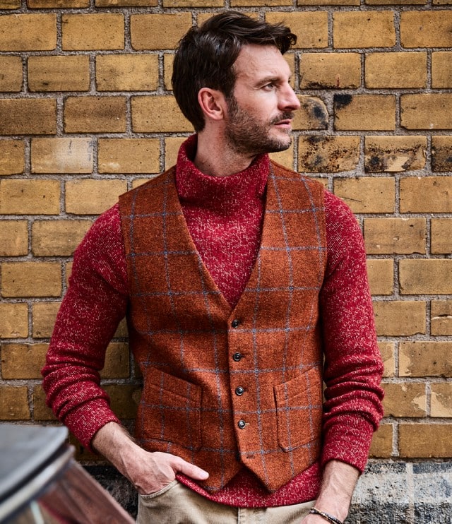 Der Mann trägt einen roten Strickrollkragenpullover unter einer orangefarbenen, karierten Wollweste und kombiniert dazu eine beigefarbene Hose – ein warmer, stilvoller Look mit herbstlichem Charakter.