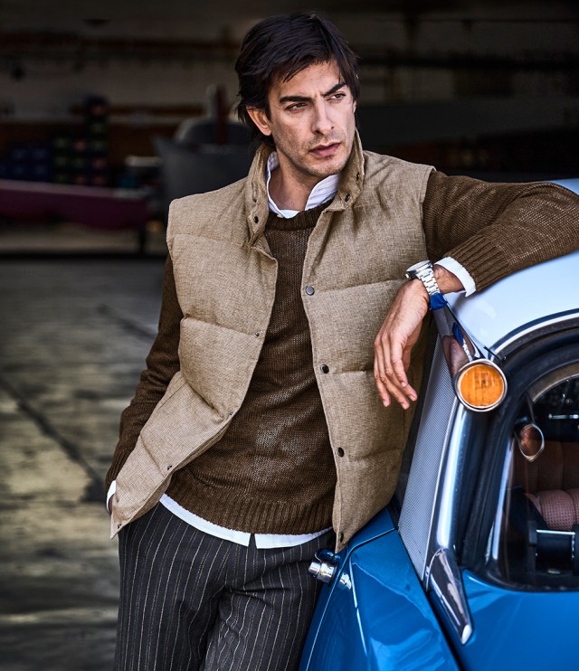 Ein Mann lehnt lässig an einem blauen Oldtimer und trägt eine beige Steppweste über einem braunen Pullover mit weißem Hemd sowie eine dunkel gestreifte Hose.