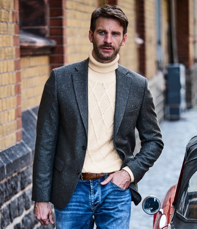 Der Mann trägt einen beigen Strickrollkragenpullover mit Rautenmuster unter einem grauen Woll-Sakko und kombiniert dazu eine blaue Jeans – ein smarter, lässig-eleganter Look mit klassischem Charme.