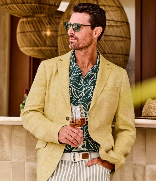 Ein Mann bekeidet mit einem sandfarbenen Sakko und einem Printhemd steht mit einem Glas Rose vor einer Strandbar.