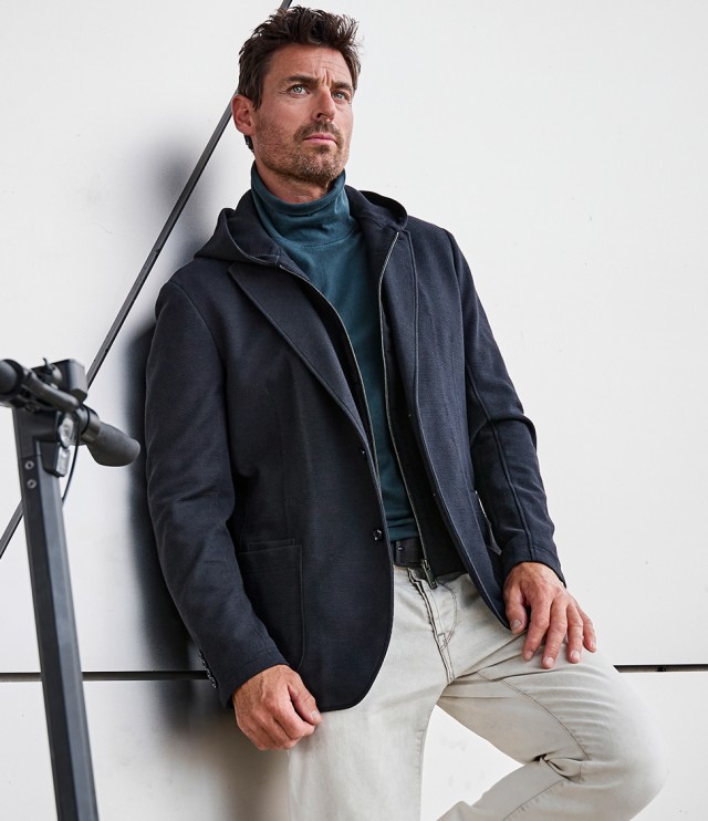 Ein Mann sitzt lässig angelehnt an einer weißen Wand, trägt eine dunkle Jacke mit Kapuze über einem petrolfarbenen Rollkragenpullover und eine helle Hose, während neben ihm ein E-Scooter steht.