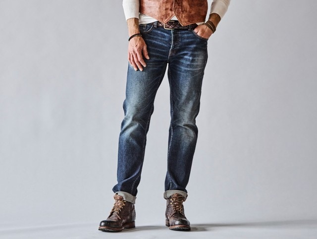 Ein Mann trägt eine ausgewaschene blaue Jeans mit umgeschlagenem Saum und braunen Lederboots, dazu ein weißes Longsleeve und eine braune Weste.