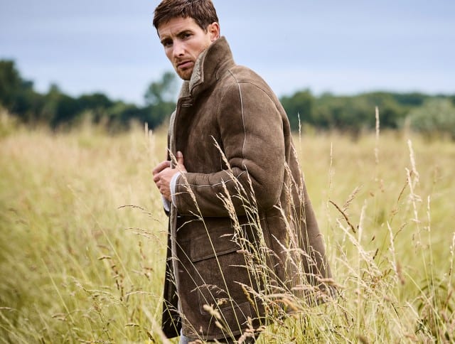 Auf dem Bild steht ein Mann in einer Wiese und trägt eine braune, warme Jacke mit hohem Kragen, während er leicht zur Seite blickt; im Hintergrund ist eine grüne Landschaft zu sehen.