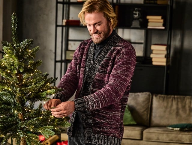 Auf dem Bild ist ein Mann zu sehen, der lächelnd einen Weihnachtsbaum mit Lichterkette schmückt; er trägt eine gestrickte, rot-schwarze Strickjacke und steht in einem gemütlich eingerichteten Wohnzimmer.