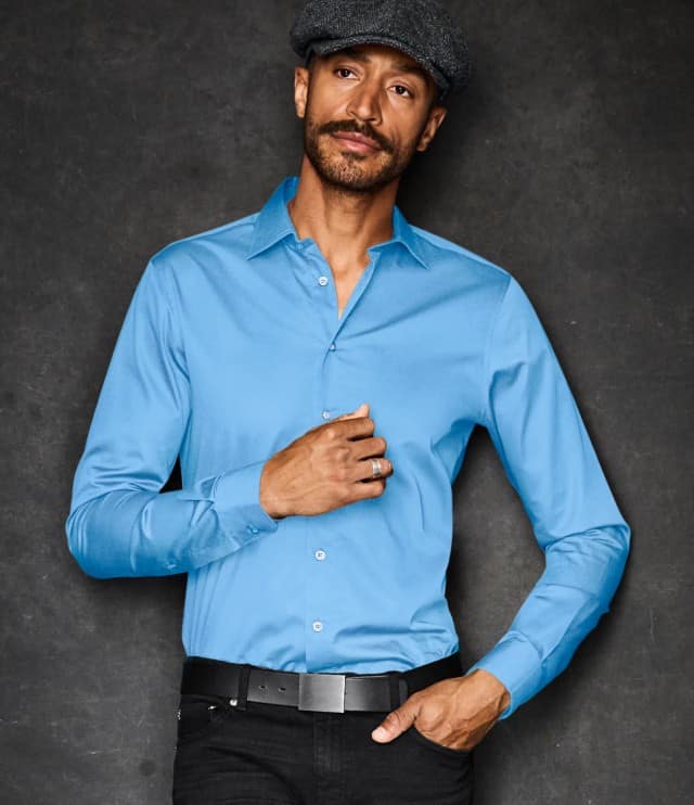 Auf dem Bild ist ein Mann zu sehen, der eine hellblaue, elegante Hemdbluse trägt und eine dunkle Schirmmütze aufhat; er steht vor einem dunklen Hintergrund und blickt leicht zur Seite.