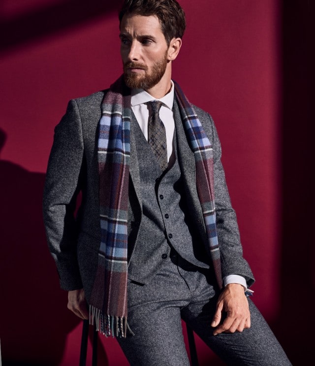 Auf dem Bild ist ein Mann zu sehen, der einen grauen Anzug mit Weste, weißem Hemd und Krawatte trägt; über den Schultern liegt ein karierter Schal, und er sitzt vor einem dunkelroten Hintergrund.