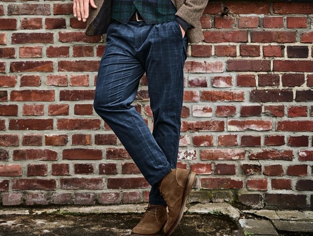 Auf dem Bild ist der Unterkörper eines Mannes zu sehen, der sich lässig vor einer Ziegelwand lehnt; er trägt blaue, karierte Stoffhosen und braune Wildlederstiefel.