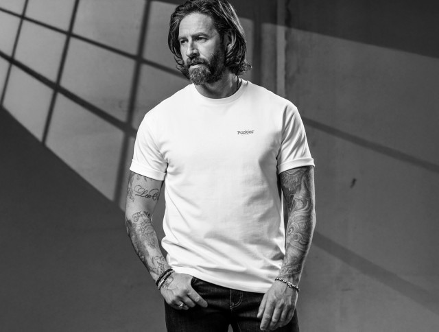 Ein Mann bekleidet mit einem weißen Shirt steht vor einer grauen Betonwand und schaut in die Ferne