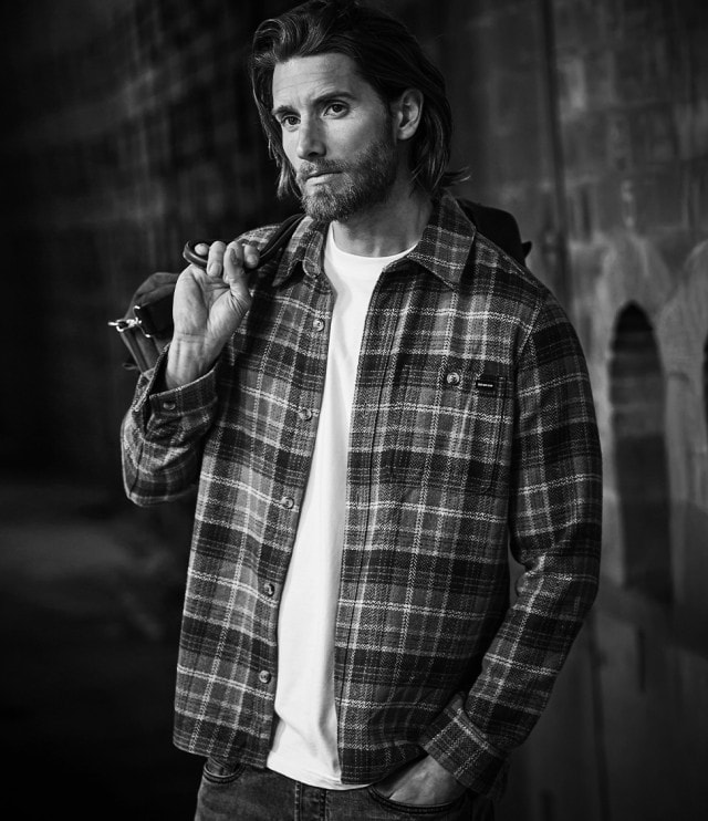 Ein Mann trägt ein kariertes Hemd über einem T-Shirt und Jeans, hat eine Tasche locker über die Schulter gelegt und blickt nachdenklich zur Seite.