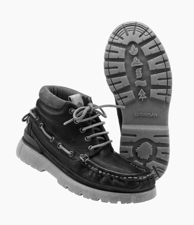 Das Bild zeigt ein Paar schwarze Lederboots mit grauen Schnürsenkeln und robuster Profilsohle, auf der verschiedene Symbole wie Feuer, Wellen und ein Baum eingeprägt sind.