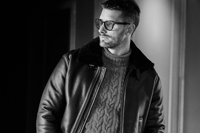 Ein Mann mit Brille trägt eine Lederjacke mit Fellkragen über einem Strickpullover und blickt zur Seite, während er im Türrahmen steht.