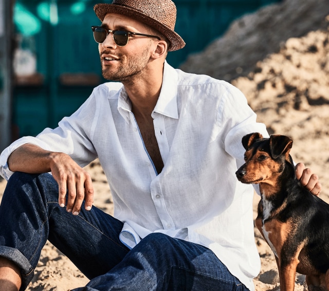 Mann mit Hut, Sonnenbrille und weißen Leinenhemd sitzt auf einem Fels neben einem kleinen Hund.