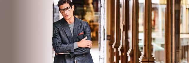 Auf dem Bild lehnt ein Mann in einem dunklen Sakko mit Brille und verschränkten Armen an einer Wand in einem elegant gestalteten Gebäude.