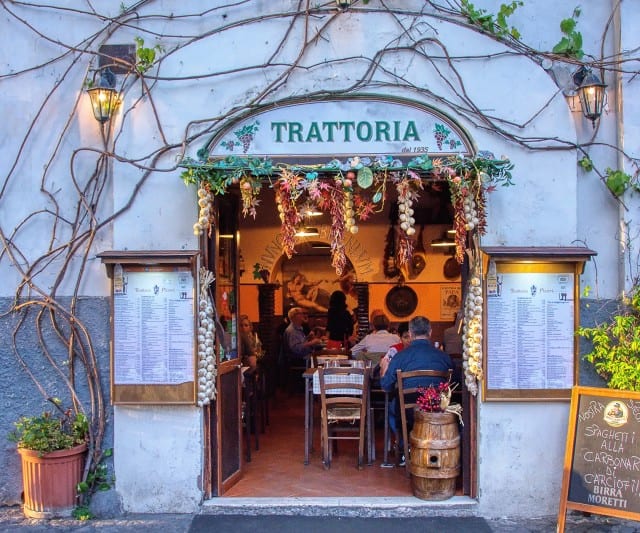 Auf dem Bild ist der Eingang einer gemütlichen Trattoria zu sehen, geschmückt mit Knoblauch, Kräutern und Lichterketten, während Gäste im Inneren an Holztischen essen.