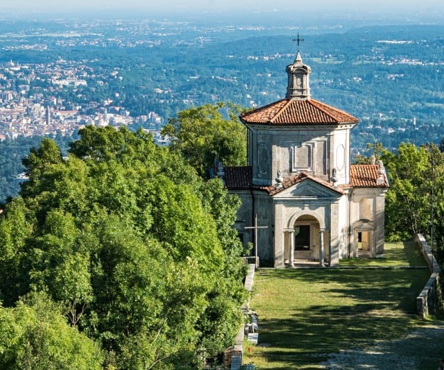 Auf dem Bild steht eine kleine historische Kapelle auf einem bewaldeten Hügel mit weitem Blick über eine Stadt und die umliegende Landschaft.