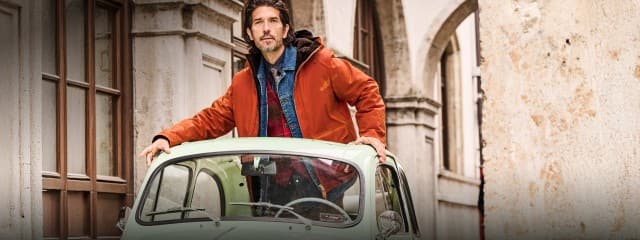 Auf dem Bild steht ein Mann in einer orangefarbenen Jacke halb aus dem Dach eines mintgrünen Oldtimers, umgeben von historischen Gebäuden.