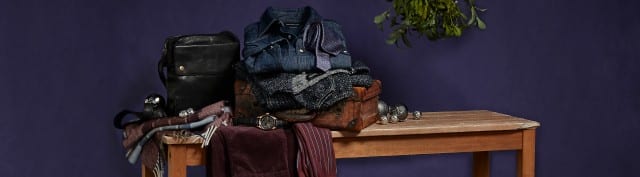 Auf dem Bild liegt eine Auswahl an Herrenbekleidung und Accessoires, darunter Hemden, Pullover, Hose, Tasche, Schal und Uhr, stilvoll auf einer Holzbank arrangiert.