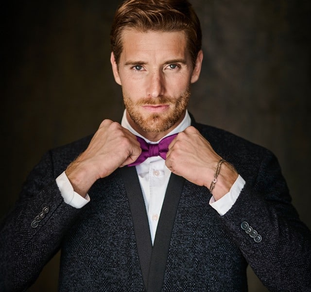 Das Bild zeigt einen Mann im dunklen Anzug und weißem Hemd, der eine violette Fliege richtet und dabei direkt und selbstbewusst in die Kamera blickt, vor einem dezent ausgeleuchteten dunklen Hintergrund.