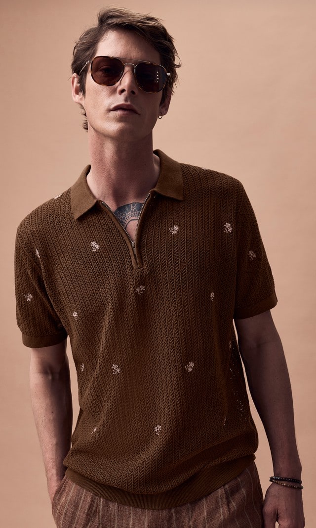 Ein Mann bekleidet mit einem gemusterten braunen Polohemd und einer Sonnebrille blickt lässig in die Kamera.