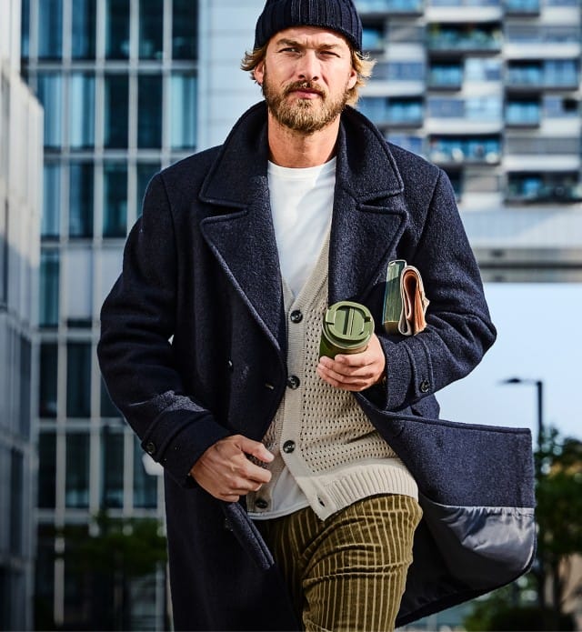 Der Mann trägt einen dunkelblauen Mantel über einem hellen Strickpullover mit weißem Shirt, kombiniert mit braunen Cordhosen und einer dunkelblauen Mütze für einen lässig-eleganten Herbstlook.