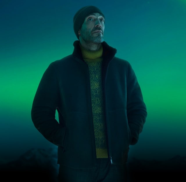 Ein Mann trägt eine Mütze, einen dunklen Mantel und einen gelben Pullover, während er mit den Händen in den Taschen vor grün leuchtenden Polarlichtern am Nachthimmel steht.