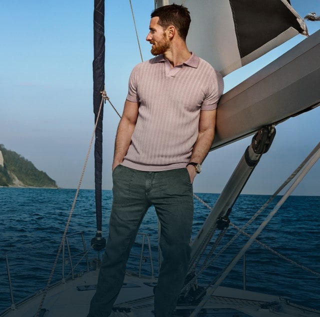 Auf dem Bild steht ein Mann entspannt auf einem Segelboot, trägt ein helles Poloshirt und eine graue Hose und blickt lächelnd zur Seite, während im Hintergrund das Meer und eine bewaldete Küste zu sehen sind.