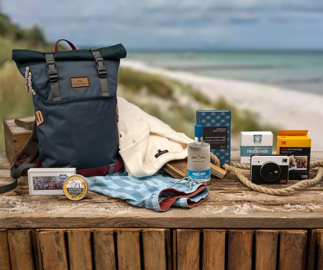 Auf dem Bild sind verschiedene Gegenstände für einen Ausflug ans Meer arrangiert, darunter ein Rucksack, Kleidung, eine Flasche Gin, ein Buch, eine Kamera, Film, Kreide, ein Abzeichen und eine Fotobox, alles auf einem Holztisch mit Strand im Hintergrund.