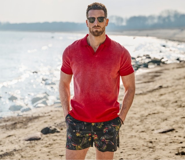 uf dem Bild steht ein Mann am Strand, der eine Sonnenbrille, ein rotes Poloshirt und gemusterte Badeshorts trägt.