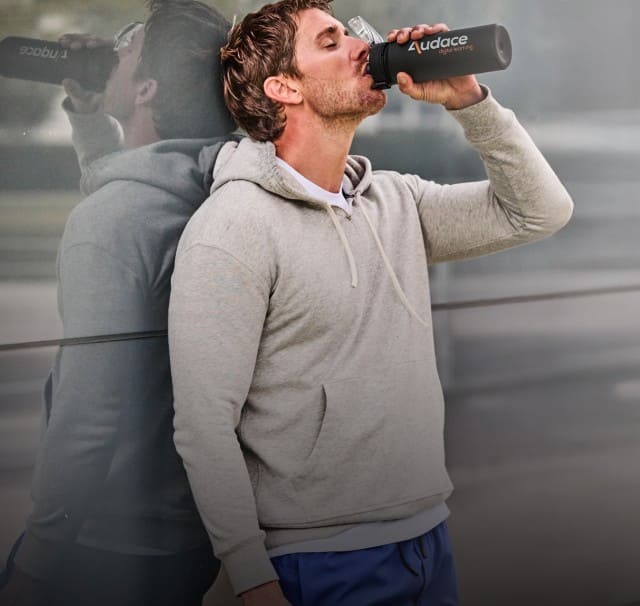 Eine Person lehnt an einer Glaswand und trinkt aus einer schwarzen Trinkflasche, während eine Spiegelung der Szene im Hintergrund sichtbar ist.