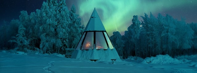 Ein schneebedecktes, zeltförmiges Holzhaus mit großen Fenstern steht inmitten eines winterlichen Waldes, während am Himmel Polarlichter zu sehen sind.
