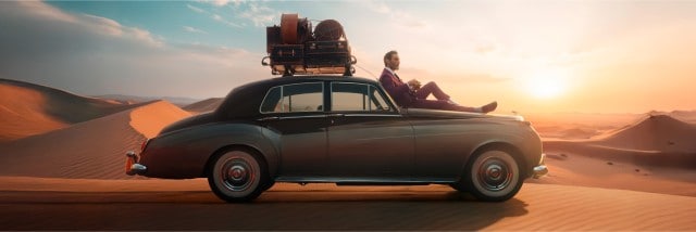 Ein Mann in elegantem Anzug sitzt entspannt auf der Motorhaube eines Oldtimers, der inmitten einer Wüstenlandschaft bei Sonnenuntergang steht, mit Gepäck auf dem Dach.