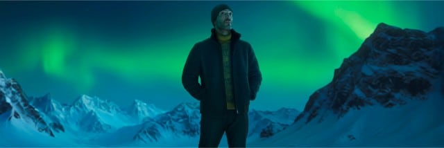 Ein Mann mit Mütze, dunkler Jacke und gelbem Pullover steht in einer verschneiten Gebirgslandschaft und blickt nach oben, während grünes Polarlicht den Himmel erleuchtet.