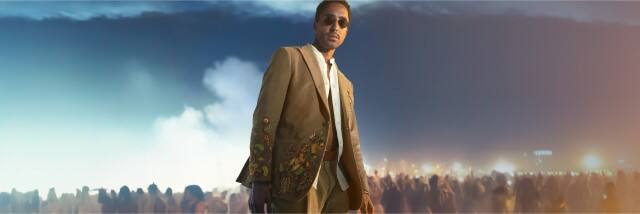 Ein Mann mit Sonnenbrille und besticktem Anzug steht vor einer Menschenmenge in nächtlicher Atmosphäre, während Lichter und Rauch den Hintergrund erhellen.