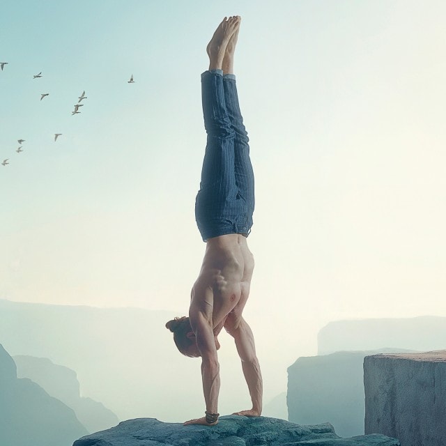 Ein Mann steht oberkörperfrei im Handstand auf einem Felsen am Rande einer Schlucht, trägt Jeans, und im Hintergrund fliegt ein Schwarm Vögel durch den Himmel.