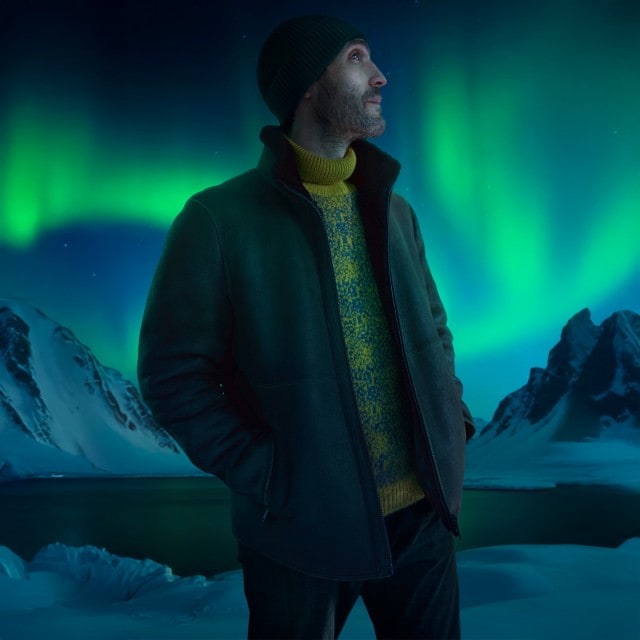 Ein Mann in warmer Kleidung mit Mütze, Jacke und Rollkragenpullover steht in einer verschneiten Landschaft und schaut nach oben, während am Himmel eindrucksvoll die Polarlichter leuchten.