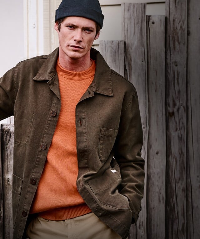 Eine Person steht lässig an einem Holzzaun und trägt eine dunkle Jacke, einen orangefarbenen Pullover sowie eine Mütze vor einer schlichten Hausfassade.