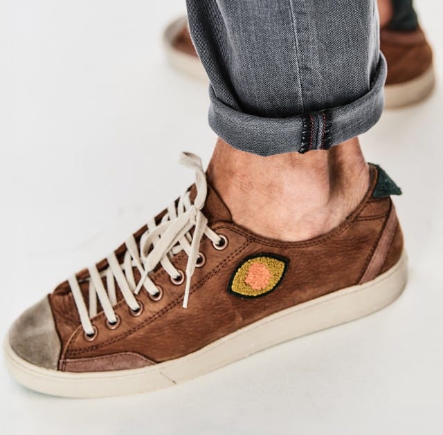 Ein geteiltes Bild zeigt links eine sitzende Person in farbiger Hose und Sneakern auf gelben Fliesen und rechts eine Nahaufnahme eines braunen Sneakerschuhs am Fuß mit hochgekrempelter Jeans und sichtbarem „SATORISAN“-Logo.