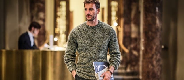 Ein Mann in einem grau melierten Strickpullover geht durch eine elegante Hotellobby und hält Zeitschriften in der Hand.