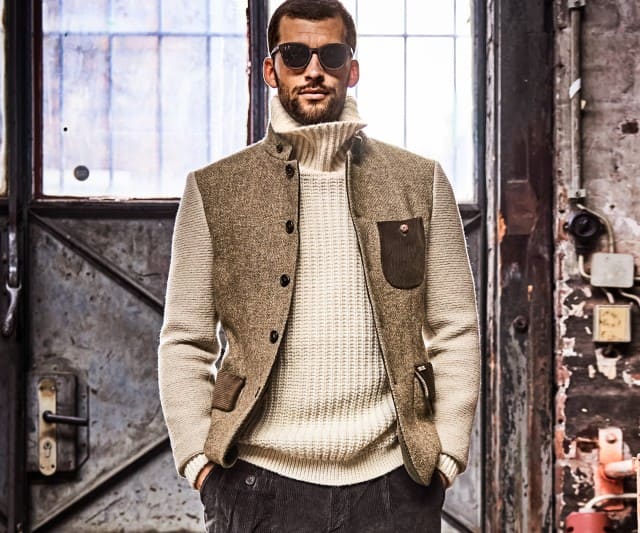 Ein Mann trägt einen dicken, cremefarbenen Rollkragenpullover und eine beigefarbene Weste, während er lässig in einer industriellen Umgebung posiert.