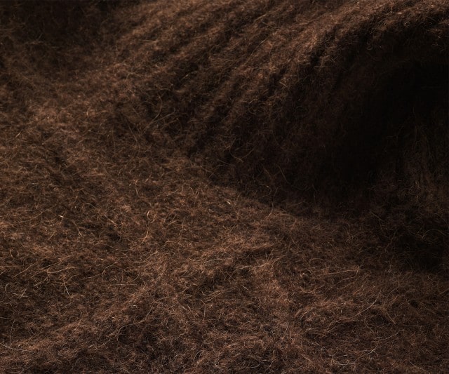 Das Bild zeigt eine Nahaufnahme von braunem, flauschigem Alpakastrick.