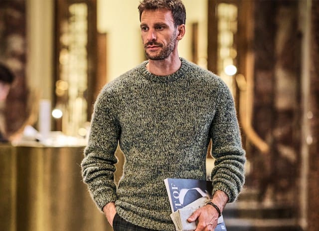 Ein Mann in einem gemusterten Wollpullover geht durch eine Hotellobby und hält eine Zeitung unter dem Arm.
