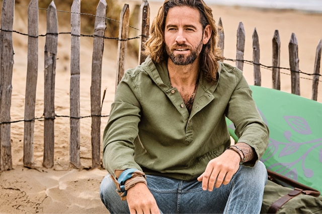 Mann in einem grünen Hemd und Jeans der vor einem Holzzaun im Sand sitzt