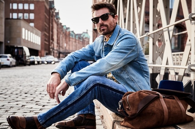 Mann mit Sonnenbrille der eine Jeansjacke und Hose trägt und auf einem Bordstein sitzt