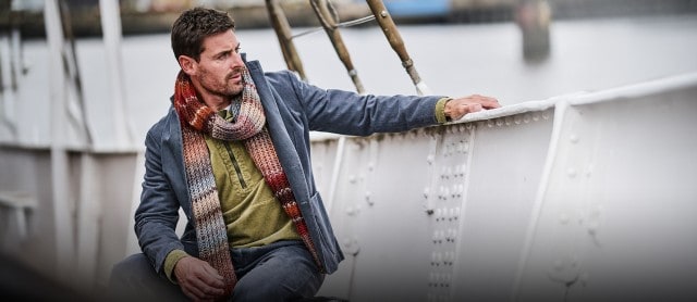 Ein Mann trägt eine olivgrüne Cord-Hemdjacke, darüber ein graues Sakko und einen bunten, gestrickten Schal in Herbstfarben