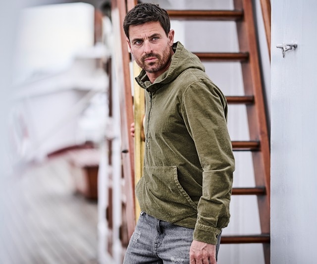 Ein Mann trägt eine olivgrüne Cordjacke mit Kapuze und Bauchtasche, kombiniert mit einer grauen Jeans.