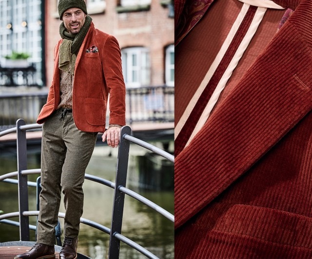 Der Mann trägt ein rostrotes Cord-Sakko mit gemustertem Hemd, olivgrüner Stoffhose mit Kordelzug, Strickschal und -mütze sowie braunen Lederschuhen – eine Mischung aus elegant und lässig.