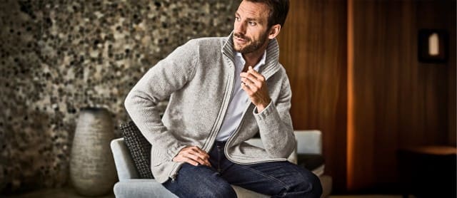 Ein Mann lehnt entspannt an einem Stuhl und trägt eine hellgraue Strickjacke.