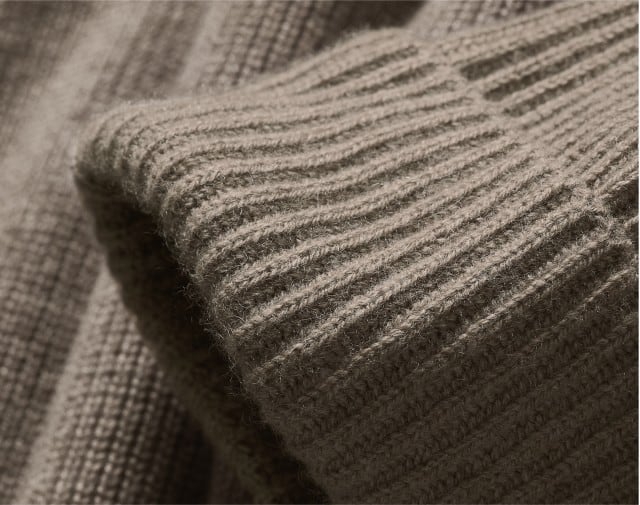 Auf dem Bild ist eine Nahaufnahme eines gerippten, flauschigen Strickstoffs in einem warmen Beigeton zu sehen, vermutlich der Ärmel eines Pullovers.